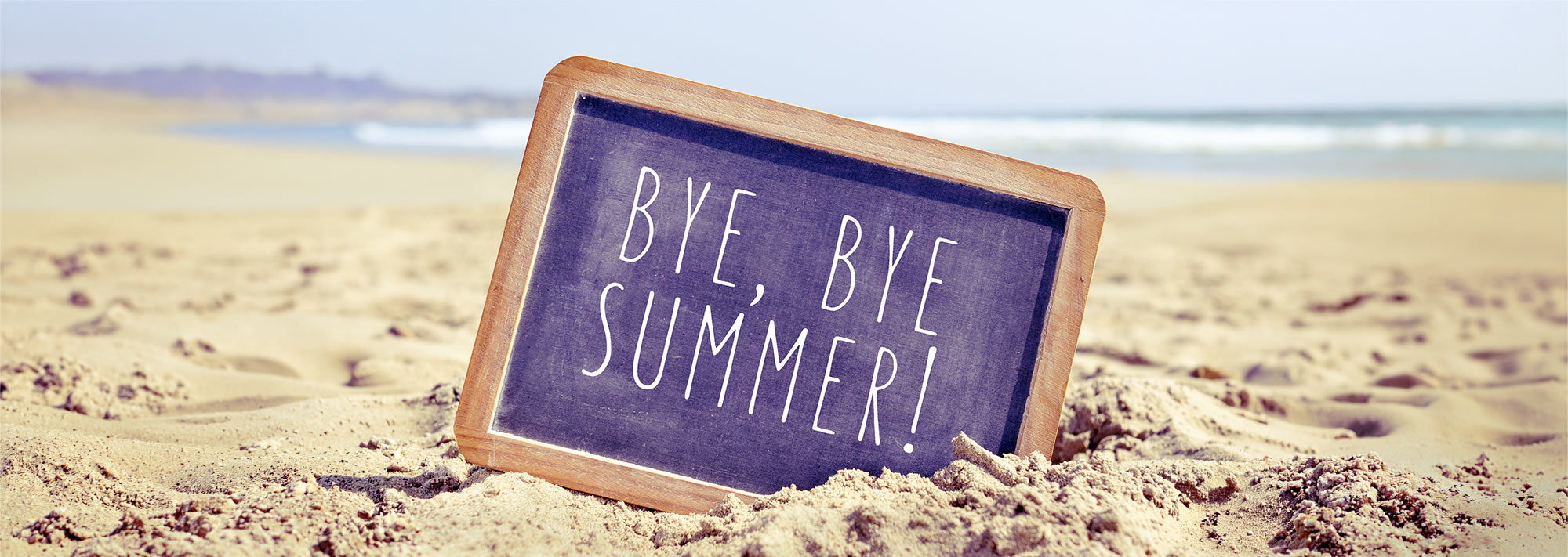 A chalkboard on a sandy beach reading "BYE, BYE SUMMER!".